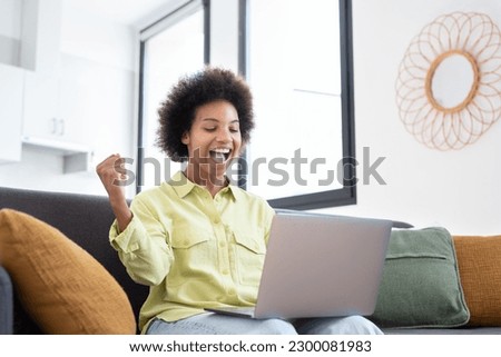 Excited cheerful young Black woman using laptop computer on sofa at home, getting good news, feeling joy, dancing with hands, singing, laughing, making winner gesture, happy to win prize Royalty-Free Stock Photo #2300081983