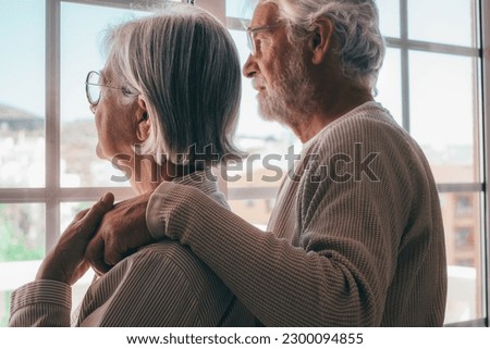 Melancholic senior family couple of Caucasian people hugging each other with love, elderly man and woman with glasses support each other in a moment of sadness looking out the window Royalty-Free Stock Photo #2300094855
