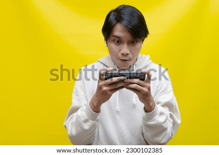 Happy expressive handsome young male gamer playing online game on smartphone isolated on yellow background