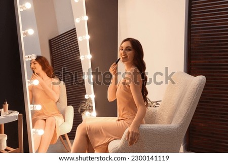 Beautiful young woman in elegant dress applying lipstick near mirror indoors