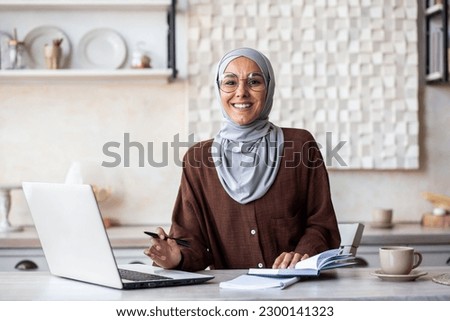 Portrait of a young Muslim female psychologist specialist in a hijab sitting at home in front of a laptop and making notes in a notebook. He consults and works online. Smiling looking at the camera.