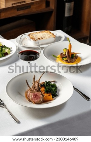 Rack of lamb grilled and cut into portions, next to it lies a piece of grilled pumpkin and fried lettuce. The food lies in a light ceramic plate on a light tablecloth, next to a glass of red wine. Royalty-Free Stock Photo #2300146409