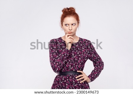 Portrait of redhead woman wearing dress holding chin and pondering idea, planning strategy, musing and solving difficult question in mind. Indoor studio shot isolated on gray background.