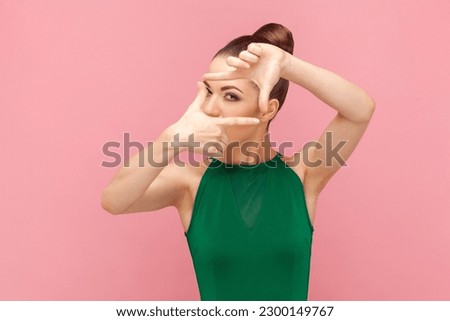 Portrait of concentrated woman photographer with bun hairstyle standing and looking at camera, making frame with fingers, wearing green dress. Indoor studio shot isolated on pink background.