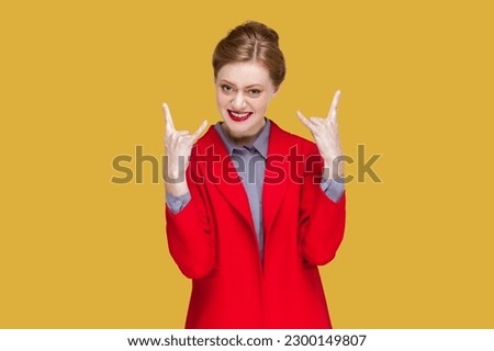 Portrait of excited crazy woman with red lips standing showing rock and roll gesture, having fun on festival, wearing red jacket. Indoor studio shot isolated on yellow background.