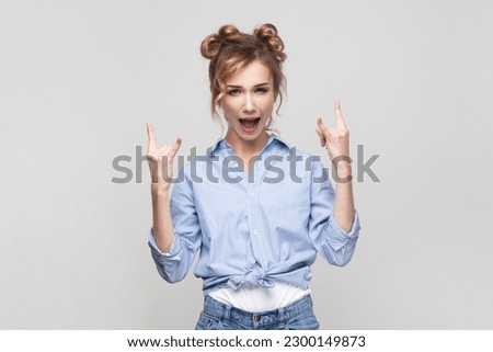 Portrait of blonde woman showing rock and roll gesture heavy metal sign enjoying favorite music on party has fun, exclaims from joy, wearing blue shirt. Indoor studio shot isolated on gray background.