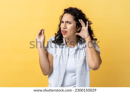 Portrait of woman with dark wavy hair holding hand near ear, listening attentively with interest private conversation, confidential talk. Indoor studio shot isolated on yellow background.