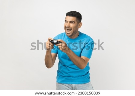 Portrait of excited extremely happy unshaven man gamer wearing blue T- shirt standing playing video games, holding joystick in hands. Indoor studio shot isolated on gray background.