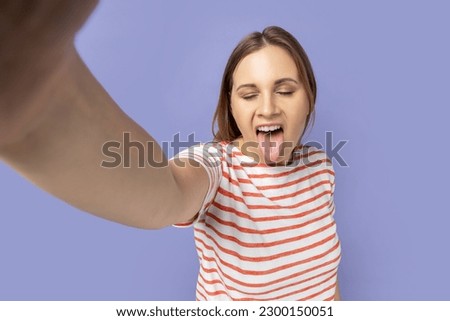 Portrait of funny childish woman in striped T-shirt taking selfie picture, point of view, showing tongue out to camera, making front selfportrait. Indoor studio shot isolated on purple background.