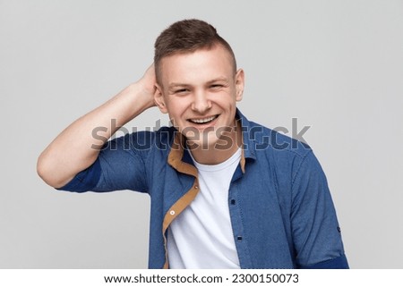 Portrait of young handsome positive friendly teenager boy wearing blue shirt looking at camera with smile, keeping hand on his head. Indoor studio shot isolated on gray background.