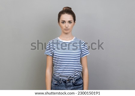 Portrait of beautiful serious bossy woman with bun hairstyle wearing striped T-shirt standing looking at camera with strict facial expression. Indoor studio shot isolated on gray background. Royalty-Free Stock Photo #2300150191