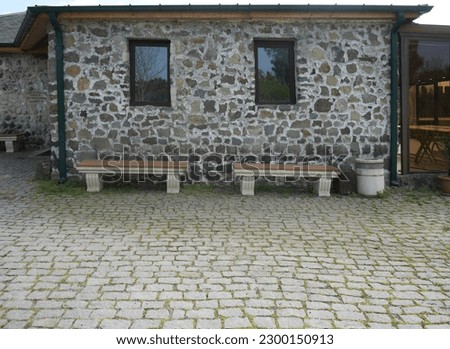 grey stone wall of house exterior with two windows and 2 benches. Historic building with wooden roof. side, front view. Cobble stone paving. 