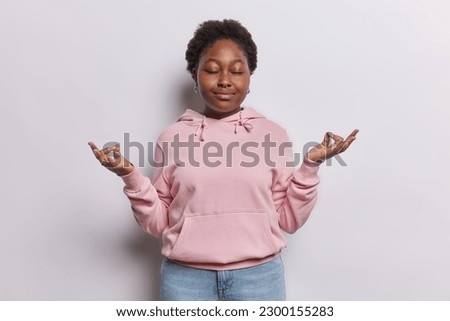 Patient calm woman makes zen nirvana gesture keeps eyes closed meditates indoor breathes deeply wears casual sweatshirt and jeans isolated over white background tries to relax for minute