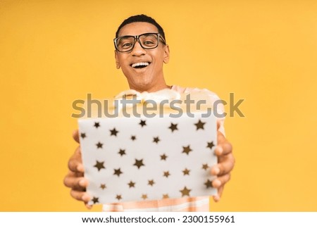 Cheerful smiling african american man holding wrapped present box. Happy young man congratulating, giving birthday gift, isolated on yellow background.