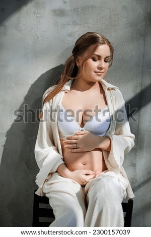 A pregnant pretty woman in a white suit and bra is sitting on a gray background. Pregnant cute woman stroking her belly and smiling. Happy period of pregnancy