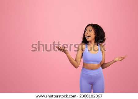 Surprised glad millennial african american lady in sportswear with open mouth hold empty space in hands isolated on pink background, studio. Sports, ad and offer, pilates, fitness and body care