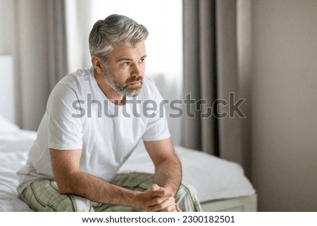 Pensive thoughtful unhappy handsome grey-haired bearded mature caucasian man wearing pajamas sitting on bed at home, looking at copy space, feeling lonely, going through divorce Royalty-Free Stock Photo #2300182501