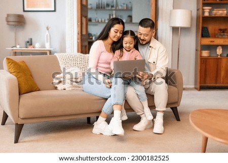 Japanese Family With Little Daughter Using Laptop Computer Watching Cartoons Online At Home, Sitting On Couch Together Spending Weekend Indoor. Internet Technology And Gadgets Lifestyle