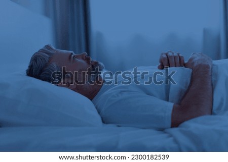 Insomnia, sleeping disorder concept. Sleepless unhappy grey-haired middle aged man wearing pajamas lying in bed late at night, watching ceiling, cant sleep, side view, copy space Royalty-Free Stock Photo #2300182539