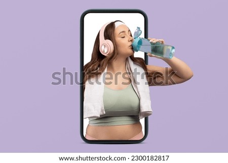 Thirsty young caucasian woman plus size in sportswear with headphones drink bottle of water on phone screen isolated on violet studio background. Music for workout, body care app, weight loss