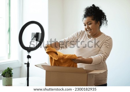 Fashion influencer happy pretty chubby young hispanic woman taking new cloth out of paper box and smiling, recommending webstore, blogger streaming while opening delivery parcel, using smartphone