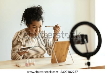 Pretty smiling curly hispanic young lady plus size makeup artist recording video for her beauty blog on smartphone, woman sitting at desk, testing brand new beauty products, eyeshadows