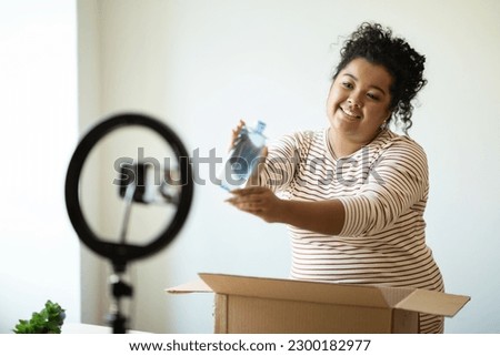Beauty blogger cute young hispanic curly woman plus size unpacking parcel at home, recording video for her blog, using blogger set phone and tripod with led lamp, showing micellar water, toner