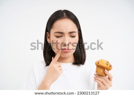 Image of pensive korean woman, looks at cupcake with thoughtful face, thinking about dessert, white background.