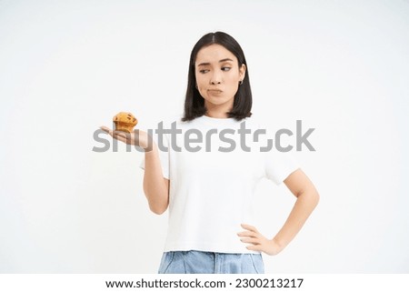 Image of korean girl, looking at cupcake and thinking, making decision, white background.
