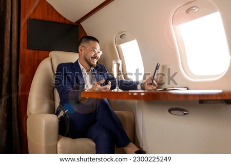 asian businessman in suit and glasses with glass of champagne flies in luxury jet and uses smartphone, korean entrepreneur films himself on the phone and communicates via video call Royalty-Free Stock Photo #2300225249
