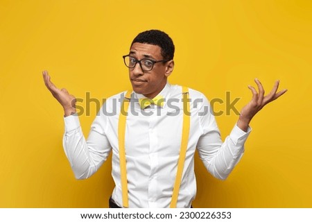 young insecure guy african american in white shirt with suspenders and bow tie spreads his arms on yellow isolated background, nerd man in glasses shrugs his shoulders Royalty-Free Stock Photo #2300226353