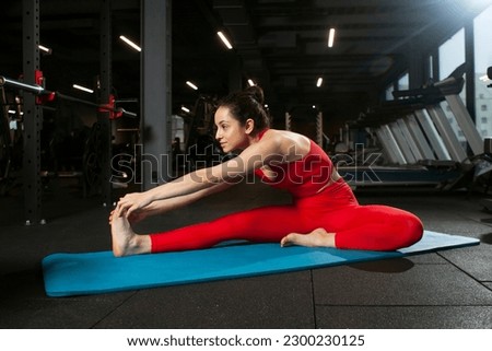 athletic woman in red sportswear sitting on yoga matte in black gym and warming up, girl doing yoga and stretching, attractive woman in fitness club doing flexibility exercise Royalty-Free Stock Photo #2300230125