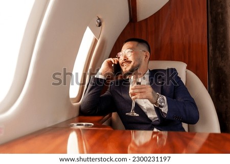 Asian businessman flies in private luxury jet and talks on the phone and drinks champagne, Korean entrepreneur in suit and glasses relaxes and communicates via mobile communication in airplane Royalty-Free Stock Photo #2300231197