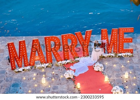 The proposal "Marry me" sentence of decorative large illuminated red letters, red carpet, champagne table, flowers and lit candles on a paving stone promenade by the sea. Concept - marriage proposal