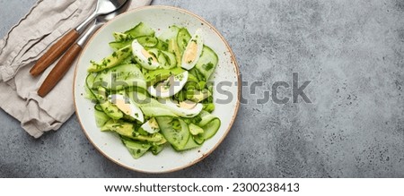 Healthy green avocado salad bowl with boiled eggs, sliced cucumbers, edamame beans, olive oil and herbs on ceramic plate top view, grey stone rustic table background. Copy space