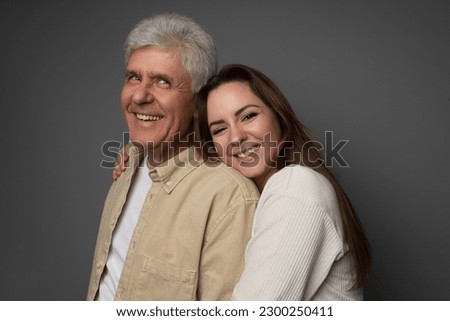 Studio portrait of a Ukrianian father in his 60s and his adult daughter in her late 20s. They are both wearing light colours and the daughter is hugging her father. The background is grey. Royalty-Free Stock Photo #2300250411