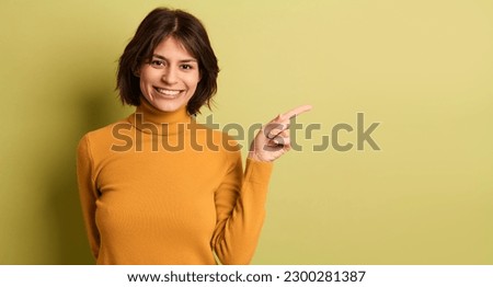 Positive young ethnic female model with dark hair in yellow turtleneck smiling brightly and looking at camera while pointing away in green studio