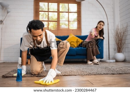 Asian young man cleaning house indoors following order from his wife. Attractive funny family, housewife housekeeper cleaner wiping on the floor for housekeeping housework or chores in house together. Royalty-Free Stock Photo #2300284467