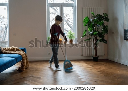 Teen boy doing chores cleaning floor in living room sweeping trash with broom to scoop. Teenager helping with household duties. Tidying up house involving children in family to maintain order at home. Royalty-Free Stock Photo #2300286329