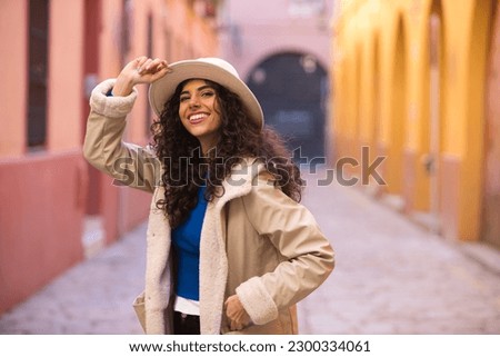 Young and beautiful Hispanic brunette woman with curly hair wearing a hat and coat for the cold walking in the city of seville while making different expressions and having fun. Royalty-Free Stock Photo #2300334061