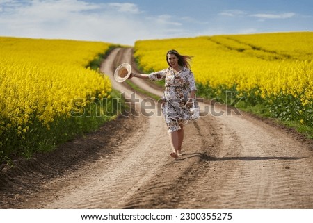 Romantic beautiful blonde hispanic young woman in a floral dress, walking barefoot on a dirt road going through a canola field