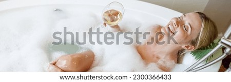 Top view on happy joyful young woman lying in bubbles in bathtub with glass of white wine.