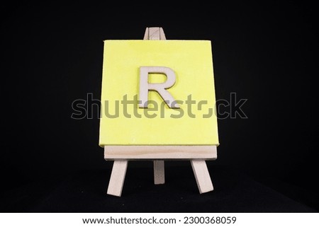 R wooden capital letter and yellow blank painting canvas resting on a miniature artists easel isolated on a black background