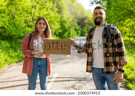 Happy couple hitchhiking on roadside trying to stop car. They are holding cardboard with inscription.