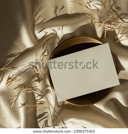 Paper card sheet with blank mockup copy space and dried grass on glossy gold silk cloth background. Aesthetic sunlight shadow silhouette. Luxury bohemian minimal invitation or business card template