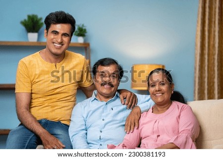 portrait shot of Happy smiling indian senior couple with adult son looking at camera while sitting on sofa at home - concept of family bonding, spending time and generation Royalty-Free Stock Photo #2300381193