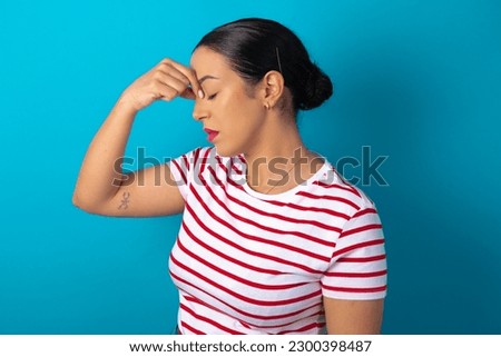 Very upset, beautiful woman wearing striped T-shirt over blue studio background touching nose between closed eyes, wants to cry, having stressful relationship or having troubles with work
