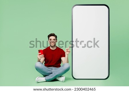 Full body fun young man he wears red t-shirt casual clothes sit point on big huge blank screen mobile cell phone with workspace area using smartphone isolated on plain pastel light green background