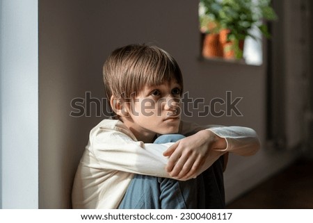 Sad worried teen boy sitting on floor embracing knees at home near window. Boring lonely frustrated teenager having life problems, troubles. Teenage crisis, bad news, melancholy, depression concept. Royalty-Free Stock Photo #2300408117