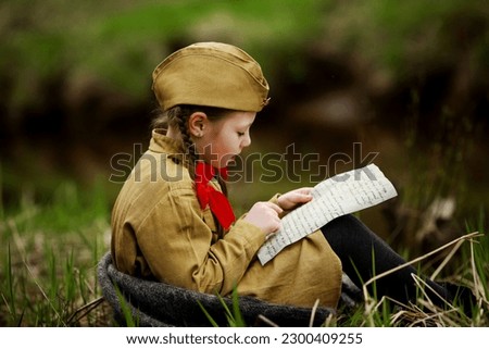 A little girl in military uniform reads an old letter from the war years sitting on the river bank. Victory Day. 9th May